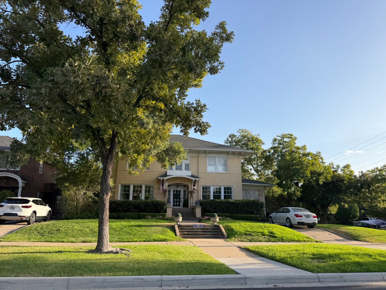 Two-story brick home behind a mature oak tree, with a landscaped lawn and broad front steps.