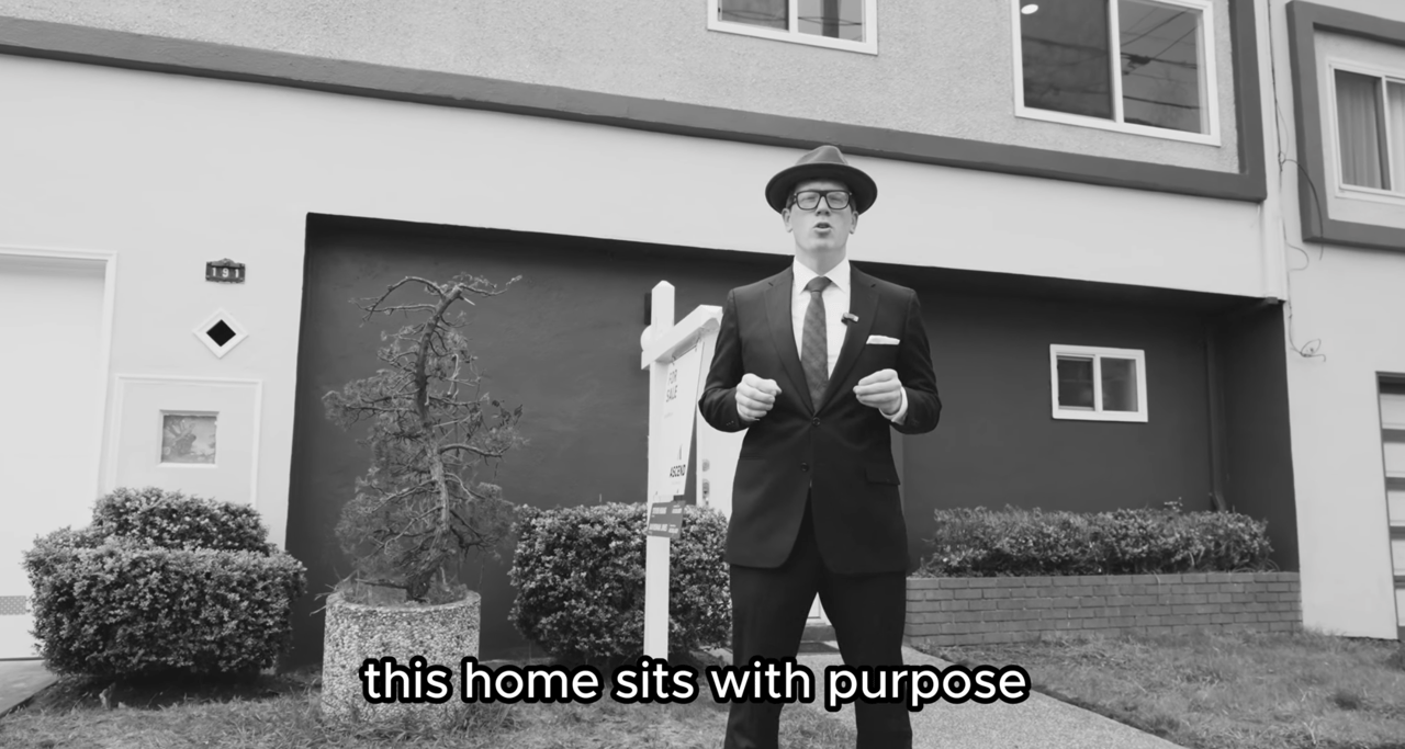 Exterior of a home with a suited presenter beside a listing sign and sculpted planter.