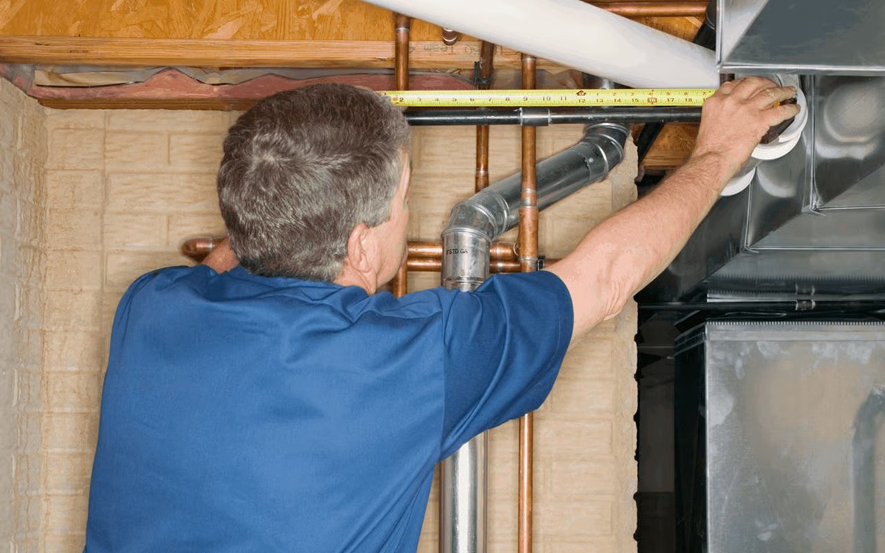 Home inspector examines furnace ductwork and pipes with a measuring tape.
