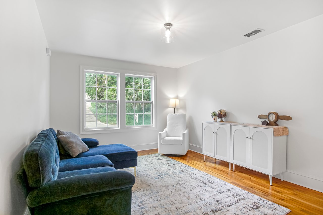 Bright living room with a blue sectional sofa, white armchair, and sideboard. Large windows let in natural light. Minimalist and cozy atmosphere.
