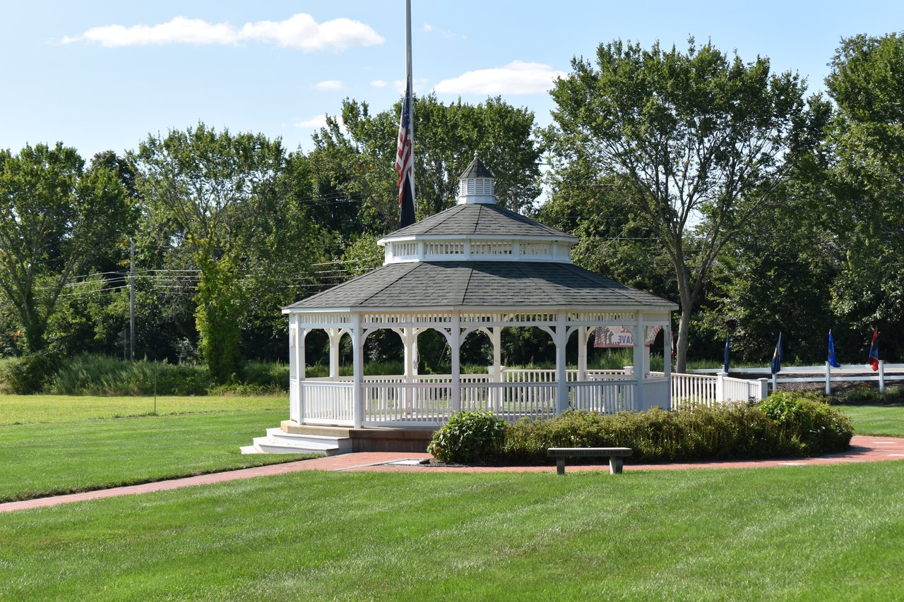 White gazebo on a green lawn in Rehoboth, with an American flag and brick path.