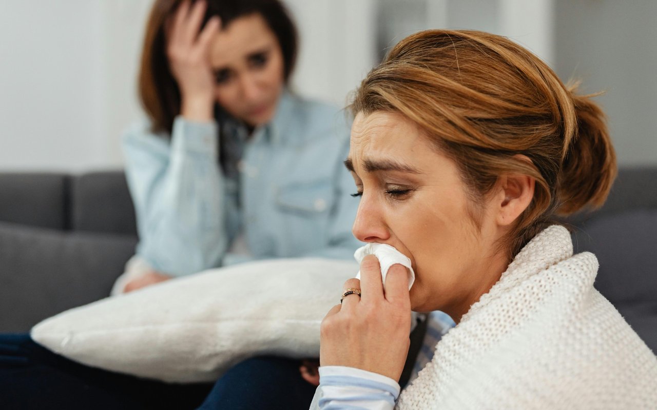 A woman wipes her nose with a tissue while another woman sits behind her.