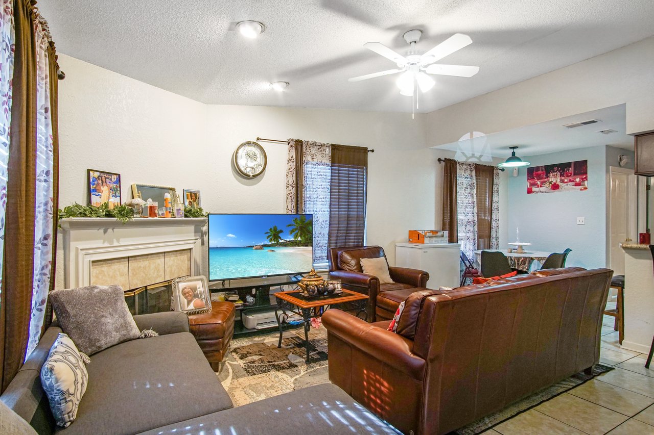 Open-concept living area at 2506 Nugget Lane in Tallahassee, Florida. The room features a tile floor, ceiling fan, and a corner fireplace with white mantel. Furnishings include a brown leather sofa set, gray sectional, and flat-screen TV. Large windows with patterned curtains bring in natural light, and the space flows into a dining nook with modern light fixture and round table.