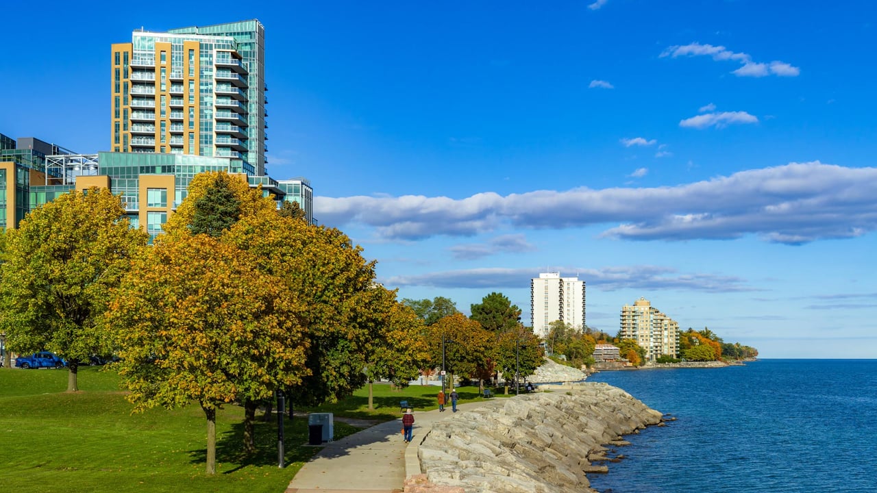 A scenic afternoon along the paved waterfront promenade near Spencer Smith Park in Burlington, Ontario, showcasing a luxury high rise condominium building next to the clear blue waters of Lake Ontario, perfect for buyers researching different Burlington neighbourhoods and considering moving to Burlington