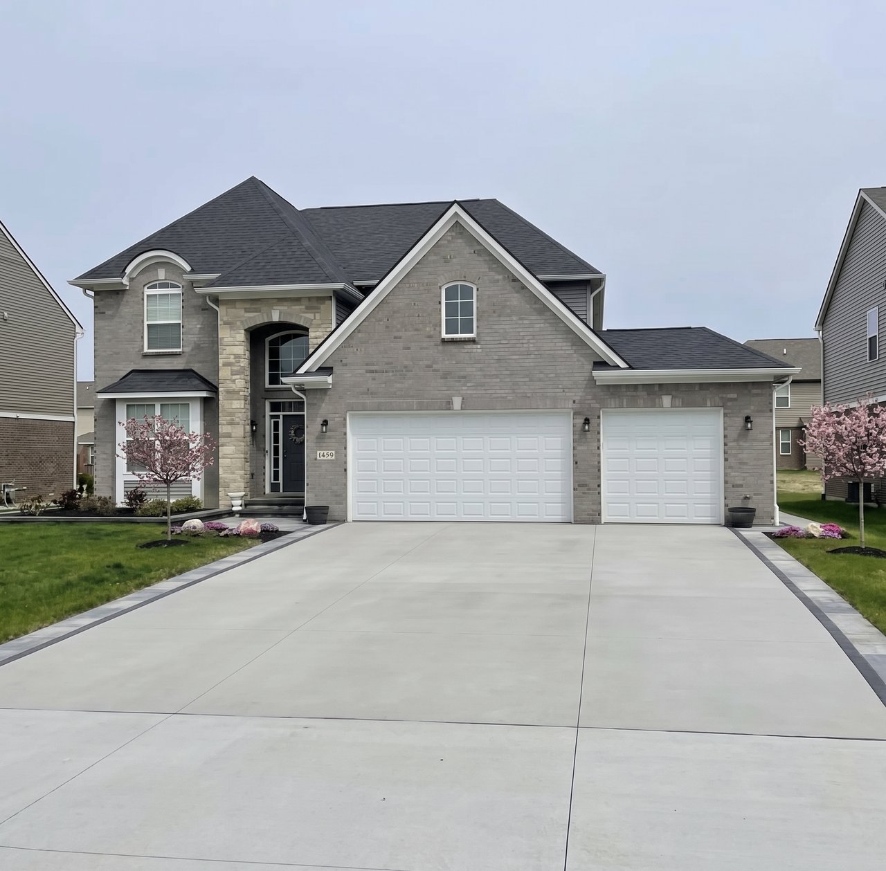 Gray brick two-story home with a three-car garage, broad driveway, and flowering pink trees.