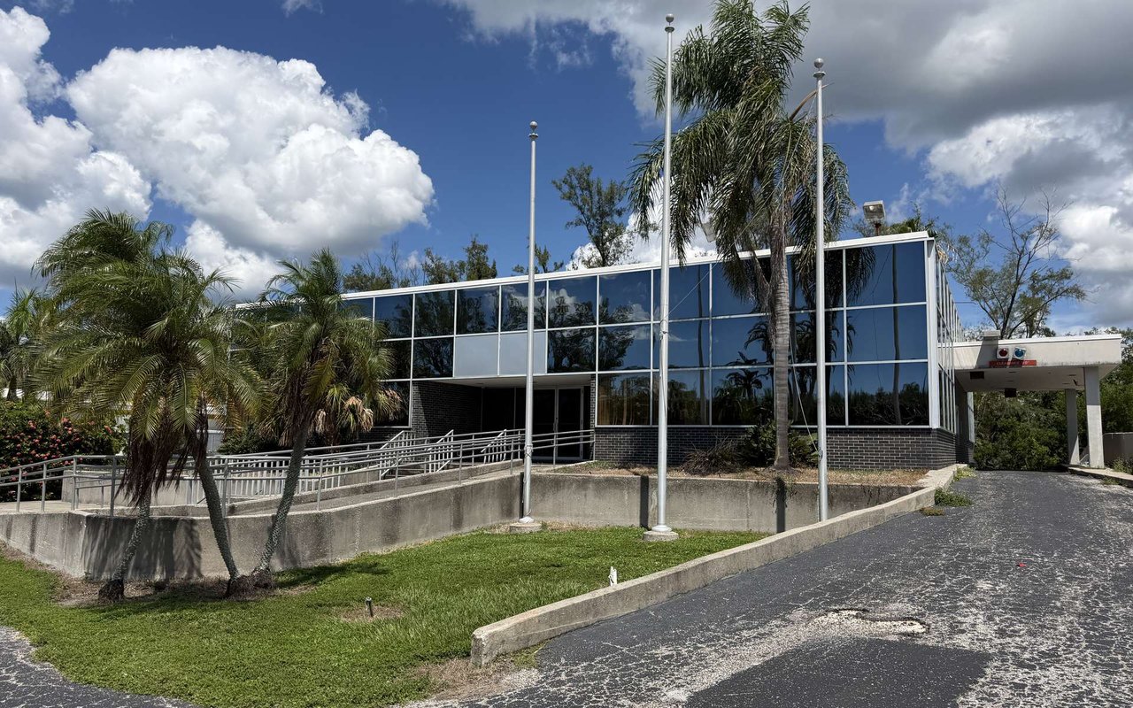 Modernist glass-fronted building with accessibility ramps, palm trees, and a covered driveway.