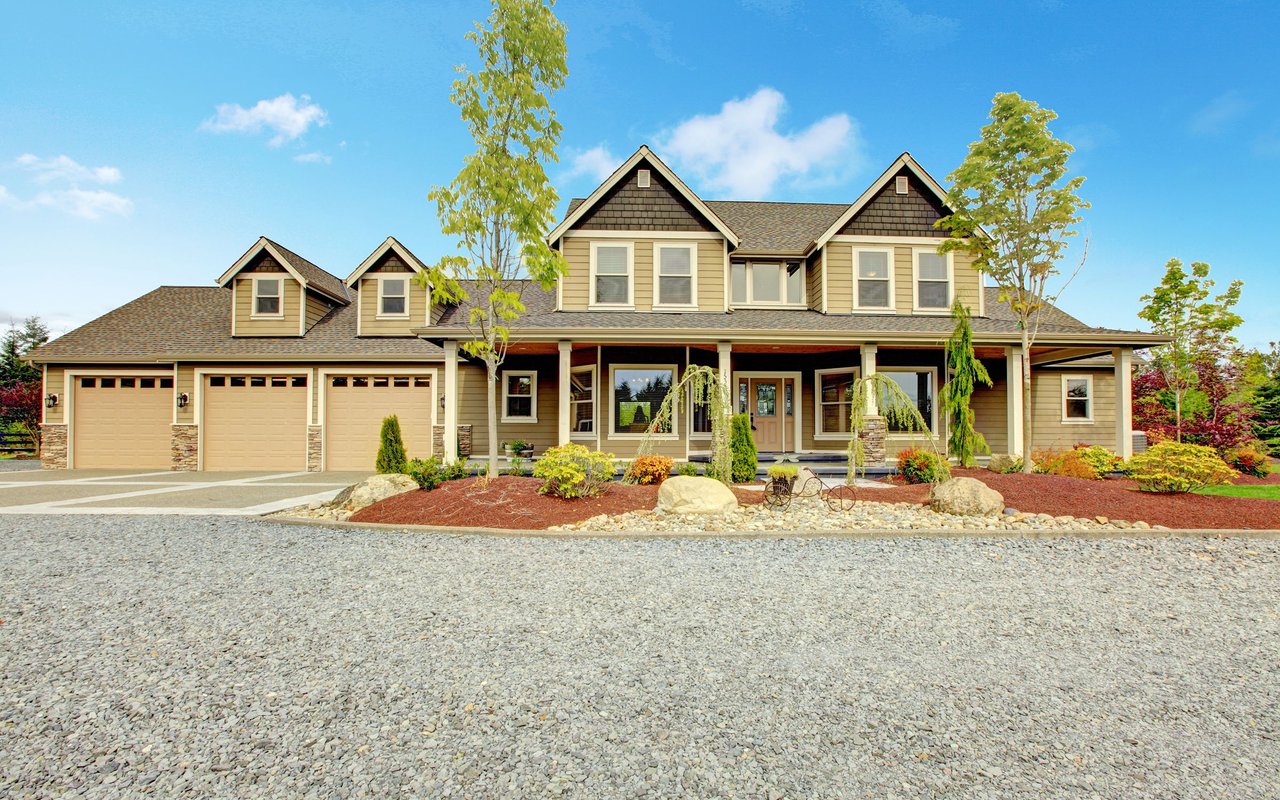 Beige two-story home with a covered porch, three garage doors, and a broad gravel driveway.