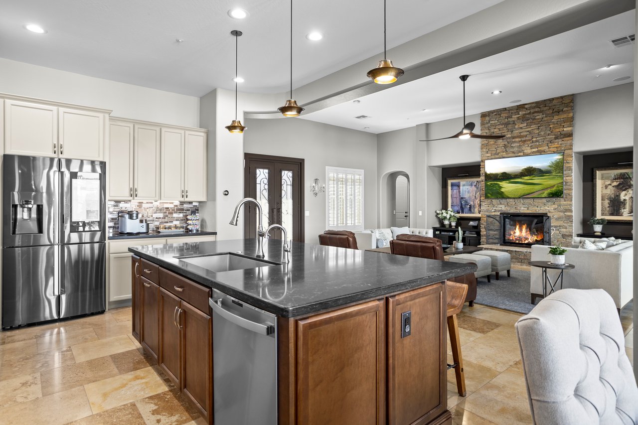 Open-concept kitchen and living room with a large island, stainless steel appliances, pendant lights, and a stone fireplace wall in the background.