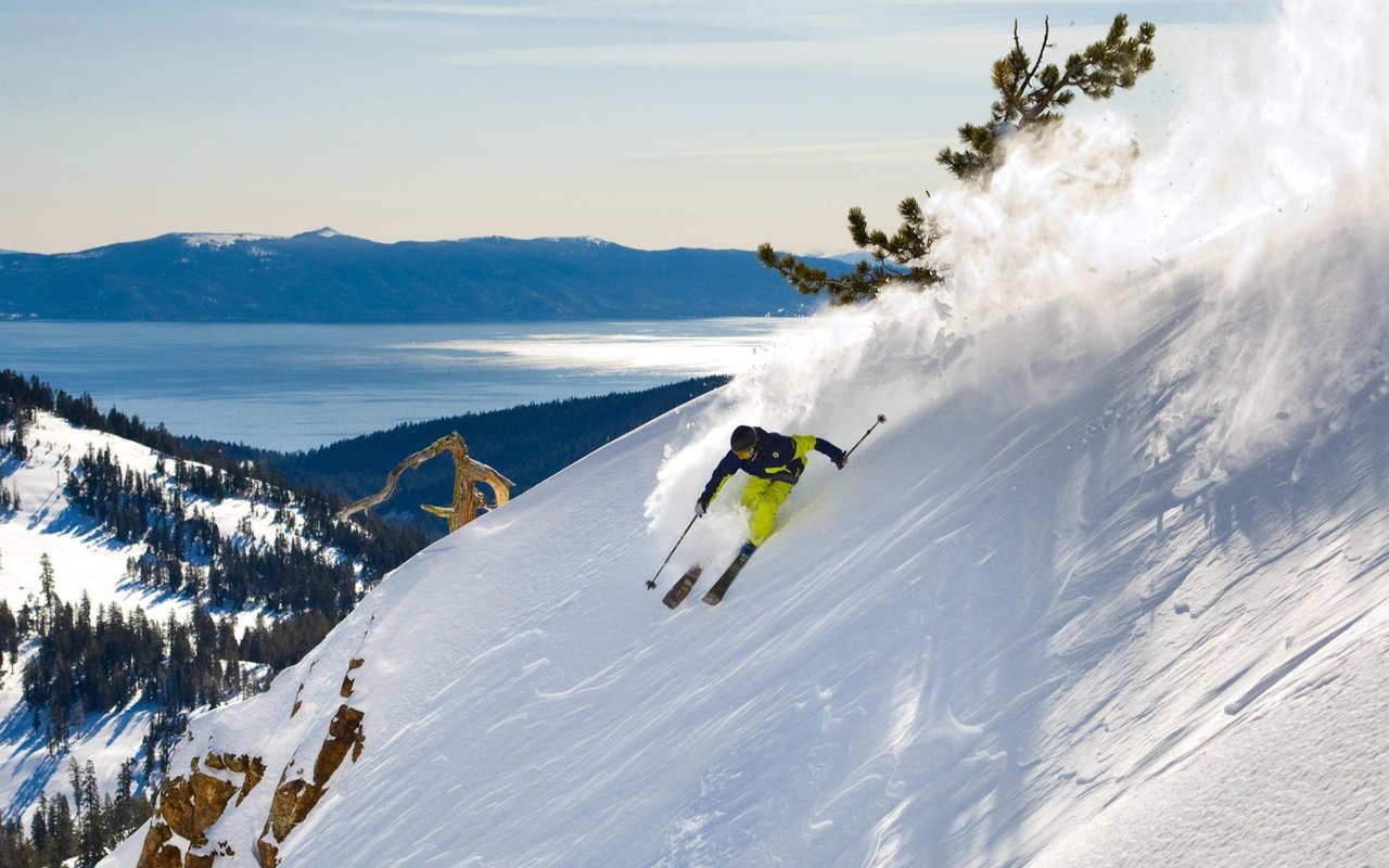 Skier carving through powder above Lake Tahoe, with forested slopes and distant mountains.