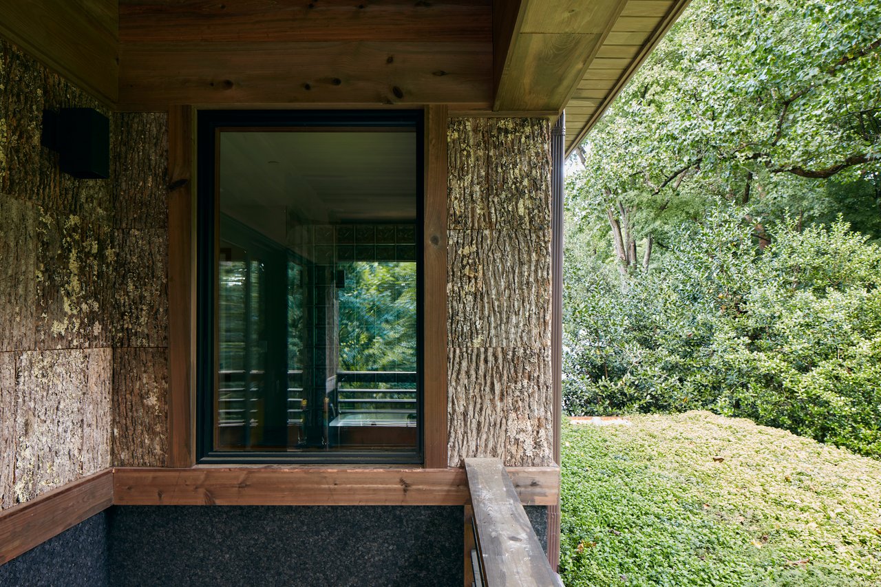 Modern home entrance with bark-textured walls, a large glass door, and a timber canopy beside lush greenery.
