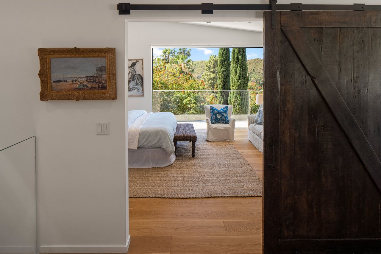 Bedroom with a dark wood barn door, woven rug, and large window overlooking trees and hillside.