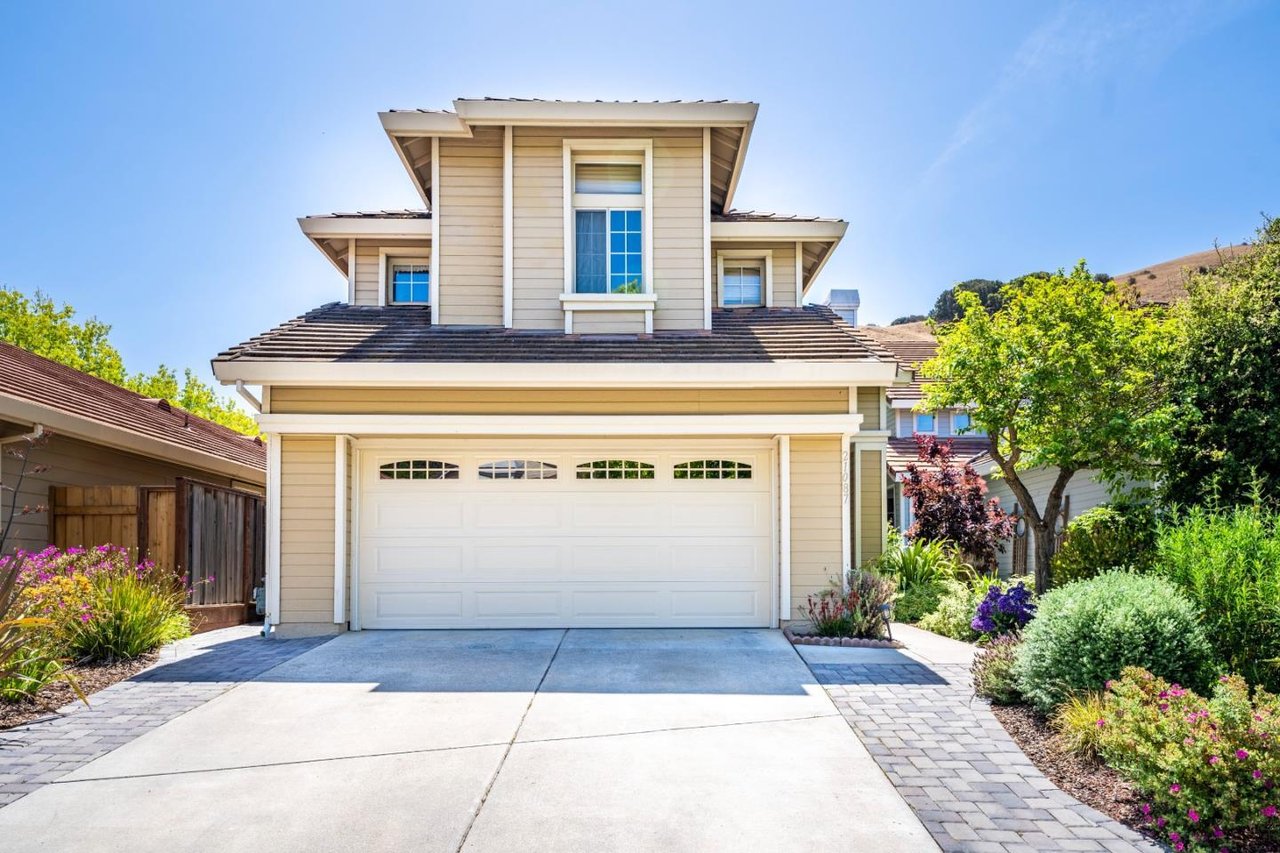 Two-story beige home with a white double garage door, wide driveway, and colorful landscaped flower beds.