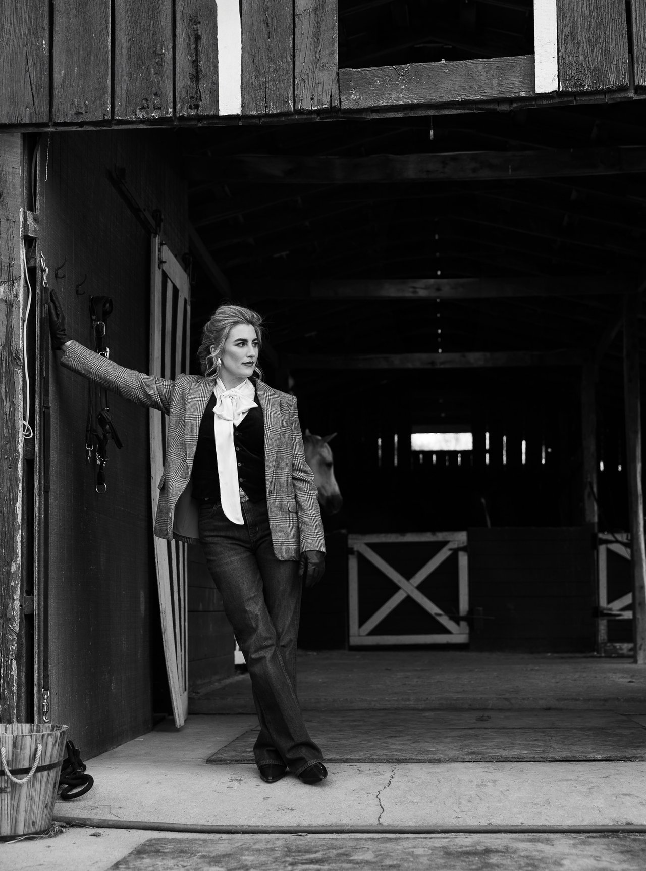 Woman in a plaid blazer posing in a wooden horse barn, with a horse partly visible behind her.