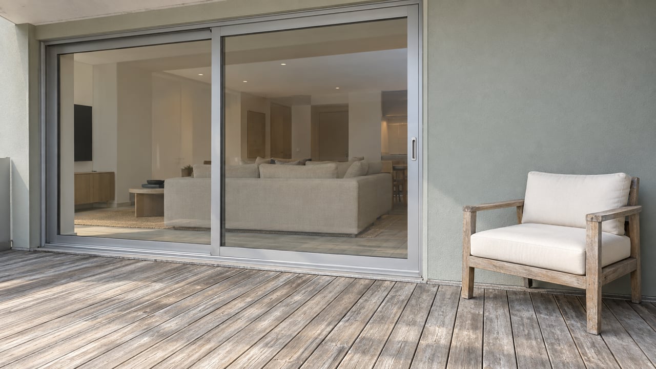 Weathered timber condominium terrace with a teak chair, pale gray-green walls and closed aluminum-framed sliding glass doors.