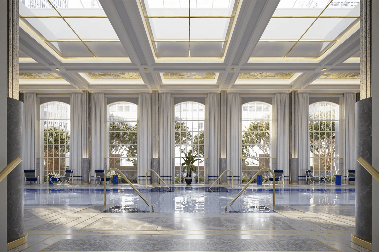 Indoor pool beneath an ornate coffered ceiling, with arched windows and marble columns.
