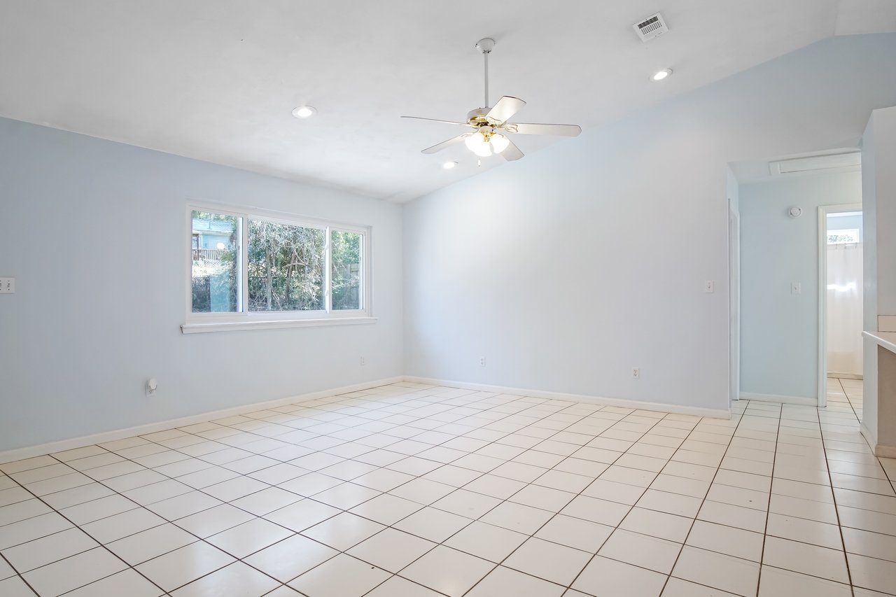 Spacious, empty room with light blue walls and white tiled floor. A ceiling fan hangs from the ceiling. Large window on the left fills the room with natural light.