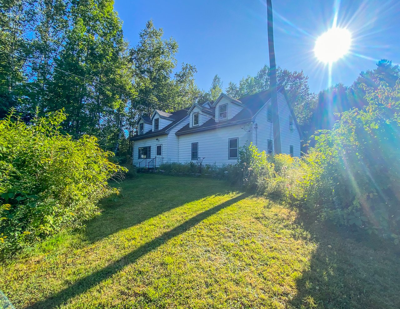 White multi-gabled house with dormer windows among trees, viewed across a sunny lawn.