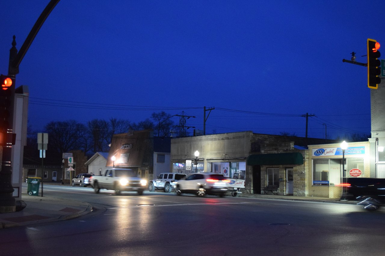 Downtown Wilmington, Illinois at dusk, with storefronts, streetlights and cars at the corner where Route 102 meets Route 53