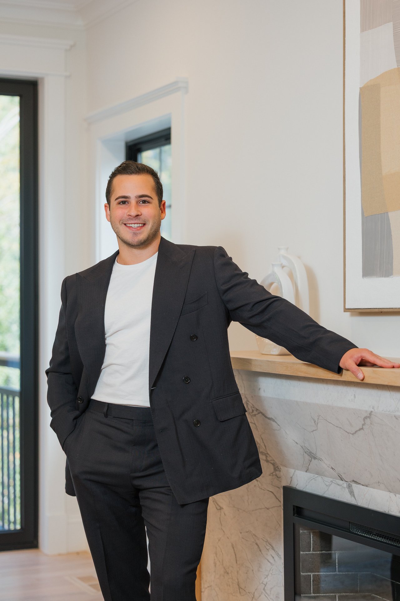 Alex MacDonald standing beside a marble fireplace in a dark suit and white shirt.