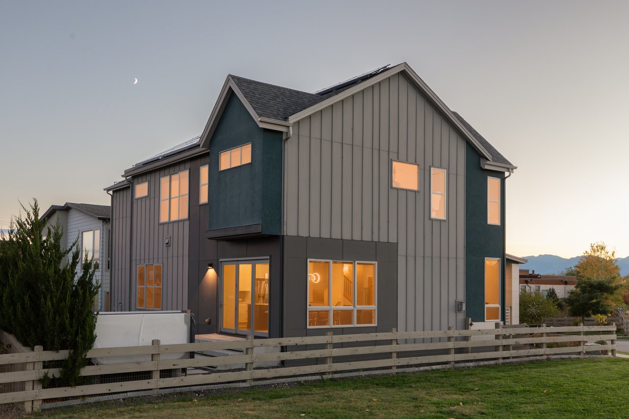 Side and rear of the home at dusk with glowing windows and a wood rail fence