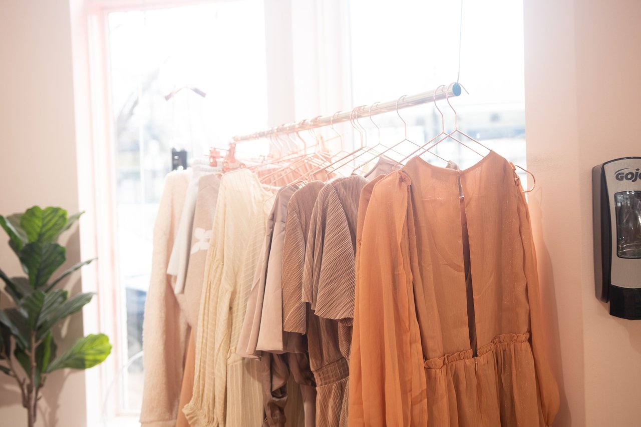 Clothing in neutral tones hangs on a rack beside a bright window and leafy plant.