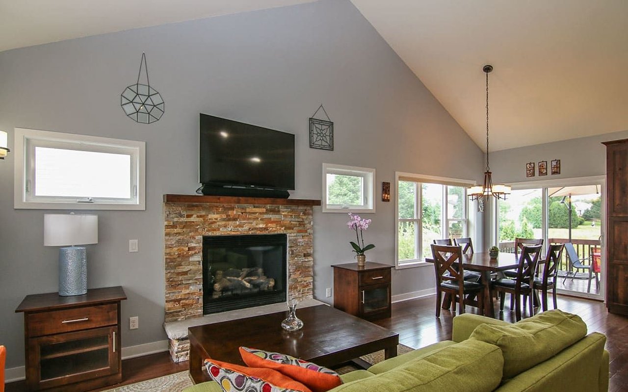 Open-plan living and dining room with a stone fireplace, vaulted ceiling, and windows overlooking a deck.