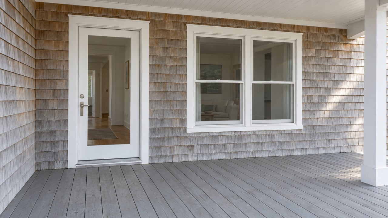 Weathered cedar-shingle home with a broad gray-painted porch, a closed glass entry door, and an off-white framed window in soft daylight.