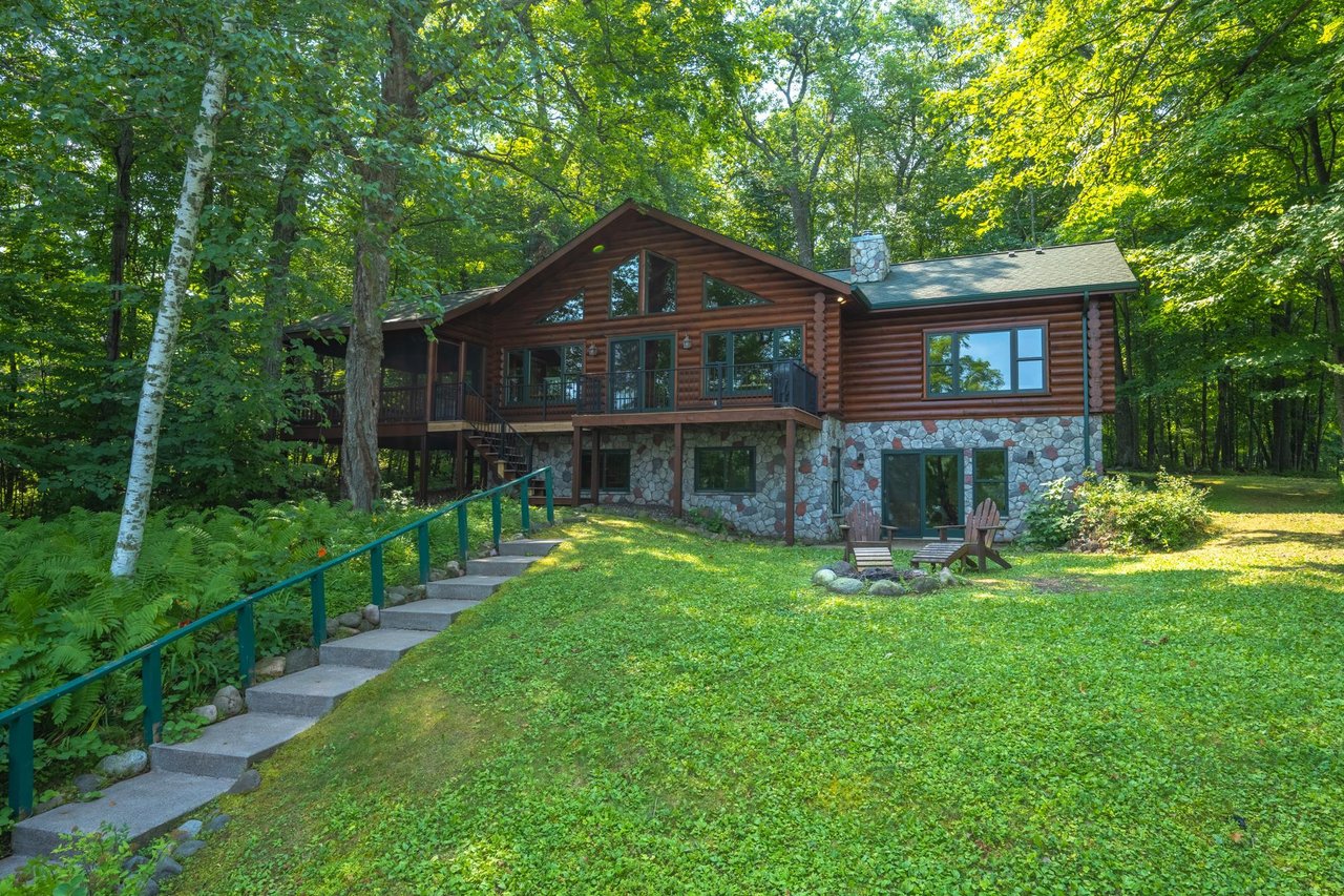Log cabin with a stone facade, raised deck, and lawn surrounded by summer trees.