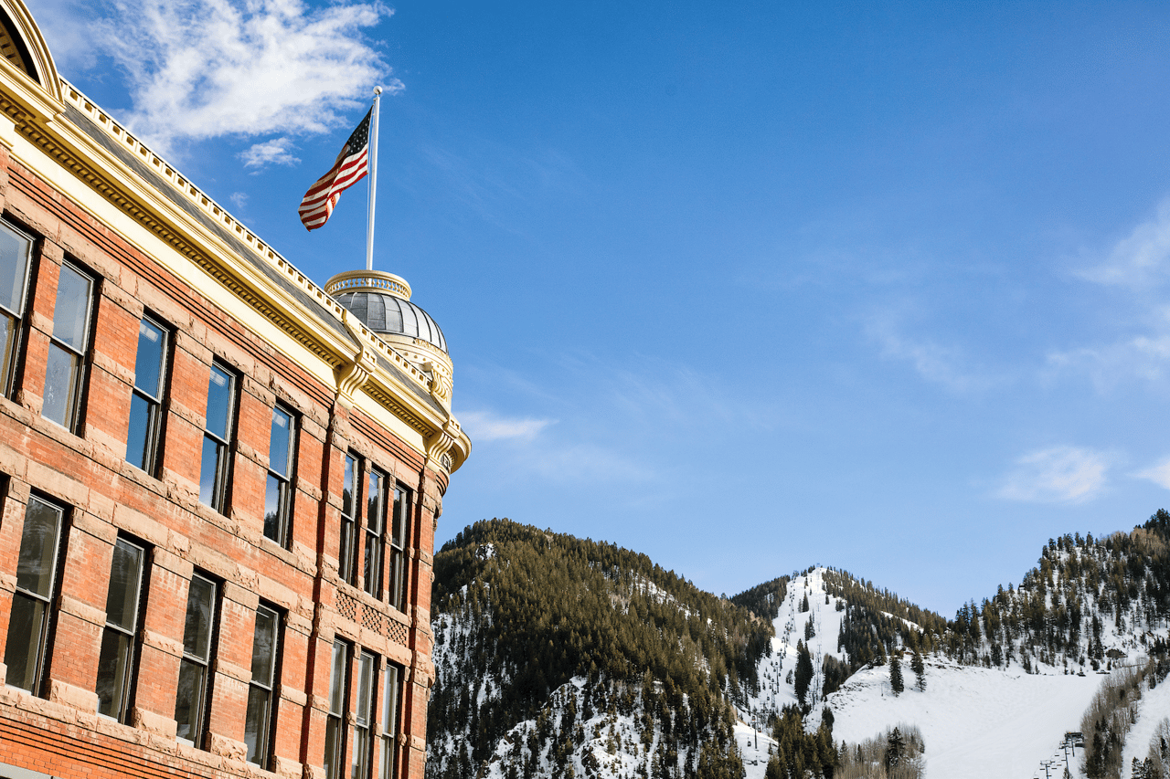 Historic brick building with American flag and distinctive rounded architecture in downtown Aspen's core neighborhood, with Aspen Mountain's ski slopes and forested peaks visible in the background against a blue sky.
