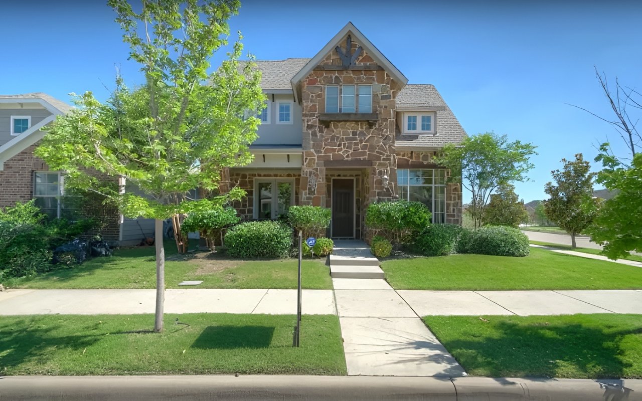 Two-story stone-front home with a peaked entry, landscaped lawn, and mature trees.