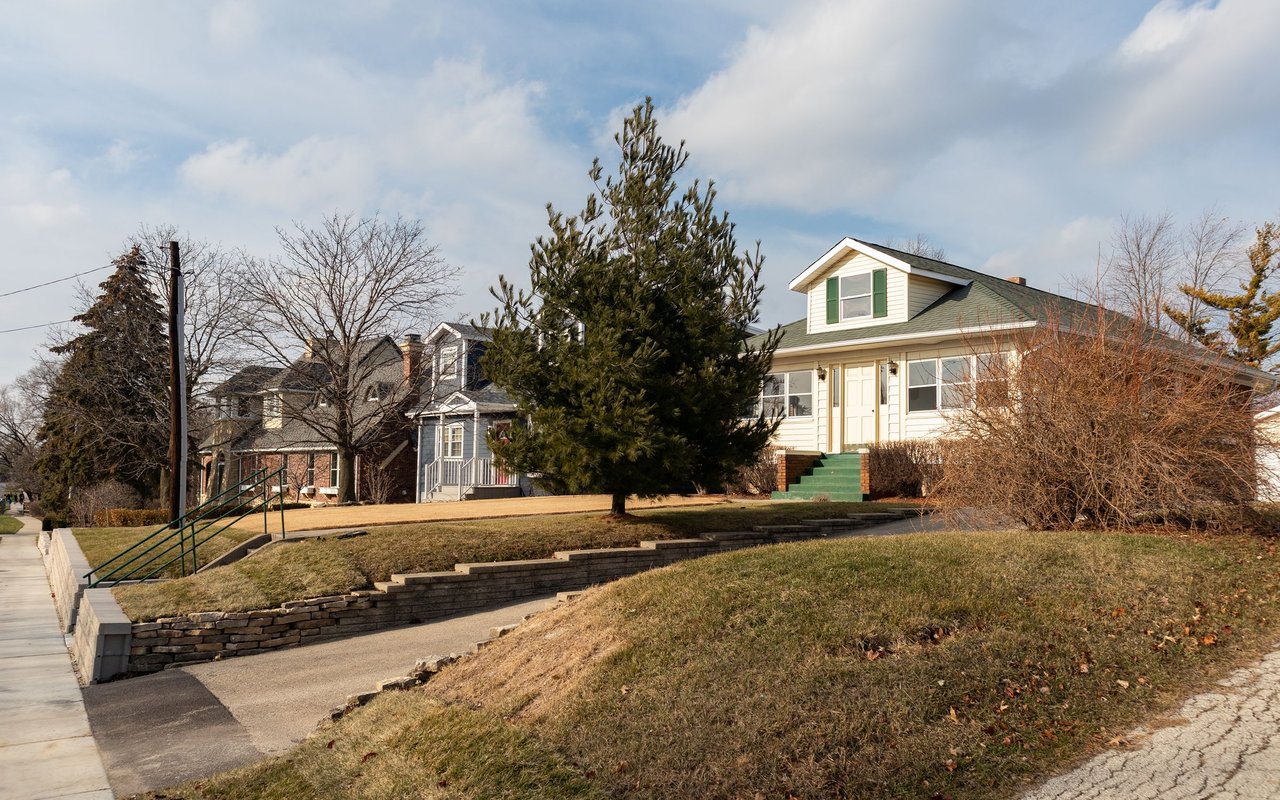 Light-colored two-story house with green shutters, raised lawn, and stone retaining walls.