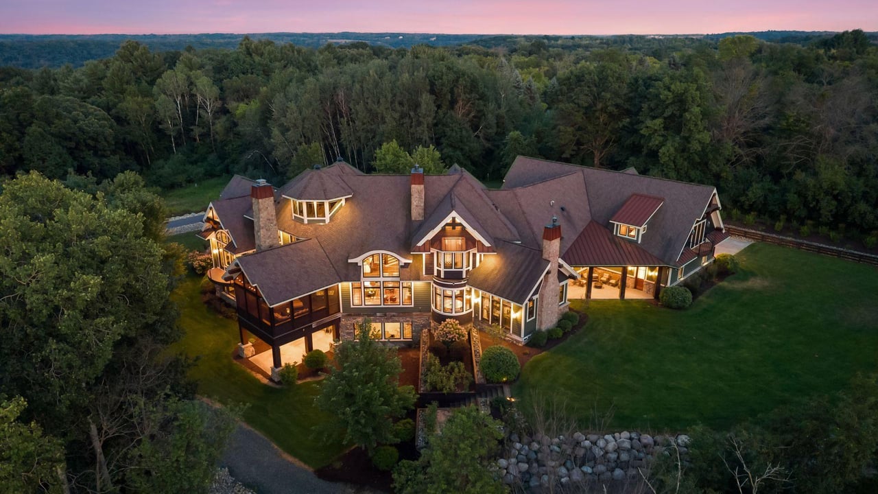 Large illuminated home amid wooded grounds at dusk, viewed from above.
