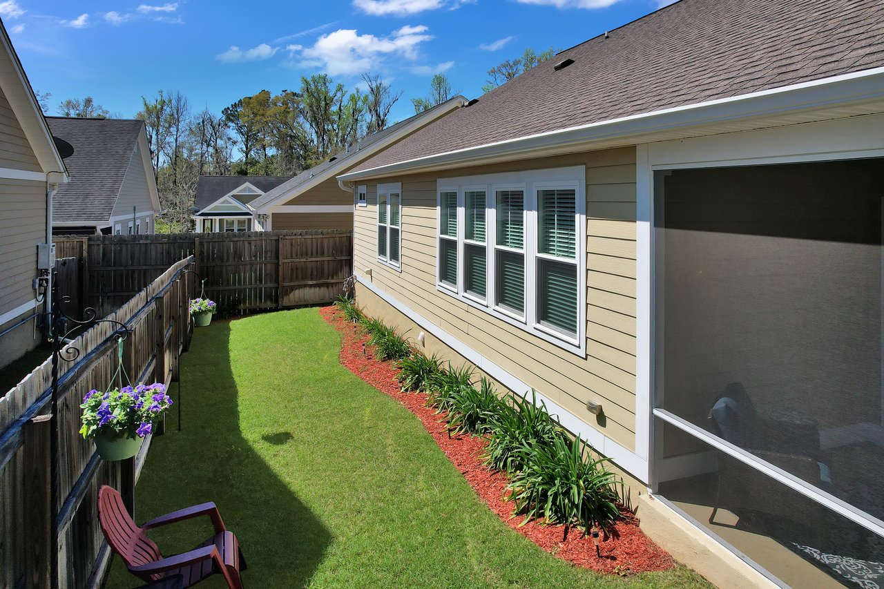 Sunny backyard with a narrow green lawn bordered by red mulch and plants. Wooden fence lined with hanging flowers and patio chair under clear blue sky.