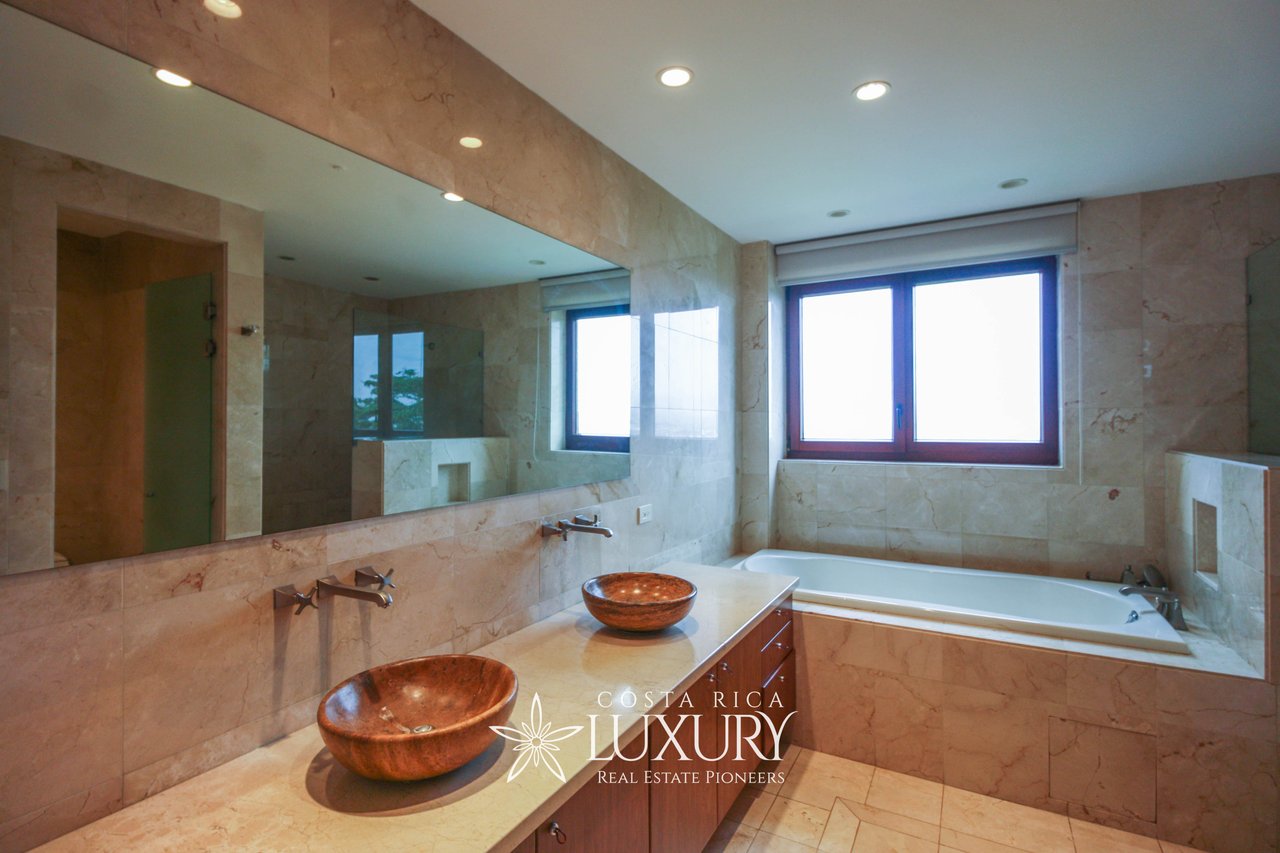 Marble bathroom with two copper vessel sinks, a long mirror, and a built-in bathtub beneath large windows.