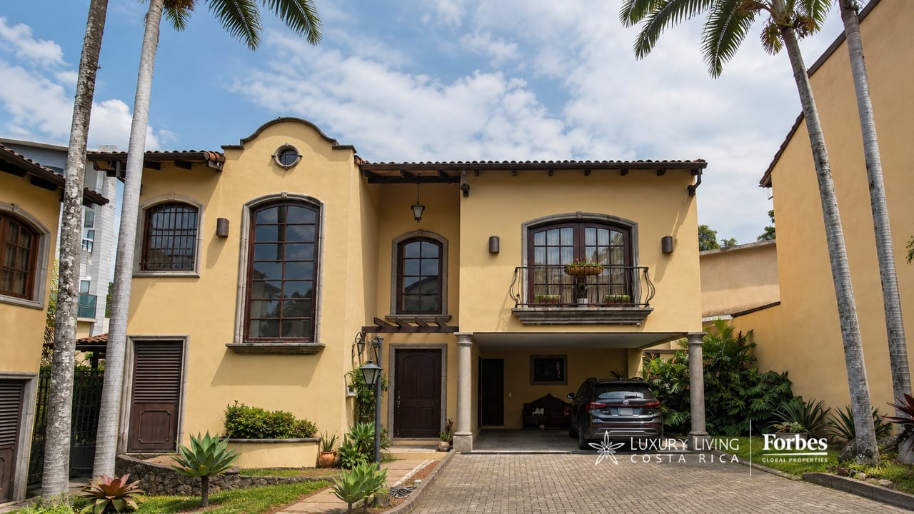 Two-story ochre house with a small balcony, palm trees, and a covered driveway.