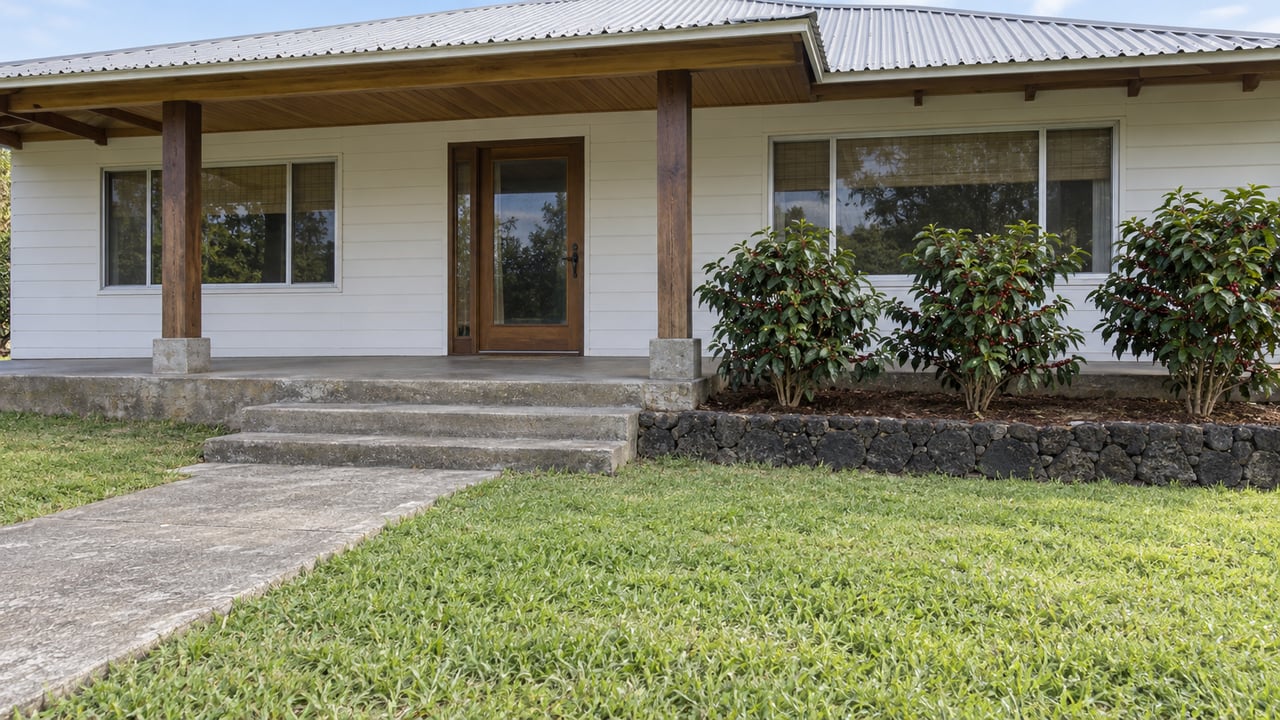 A modest wood-sided home with a corrugated roof, shaded porch, concrete entry path, and three coffee shrubs behind lava-stone edging.