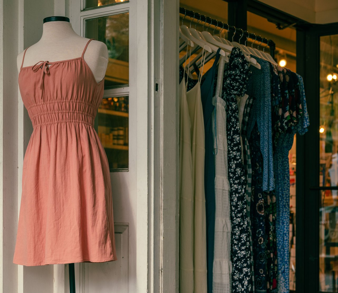 Boutique window display with a mannequin in a coral-pink dress beside a rack of dresses.