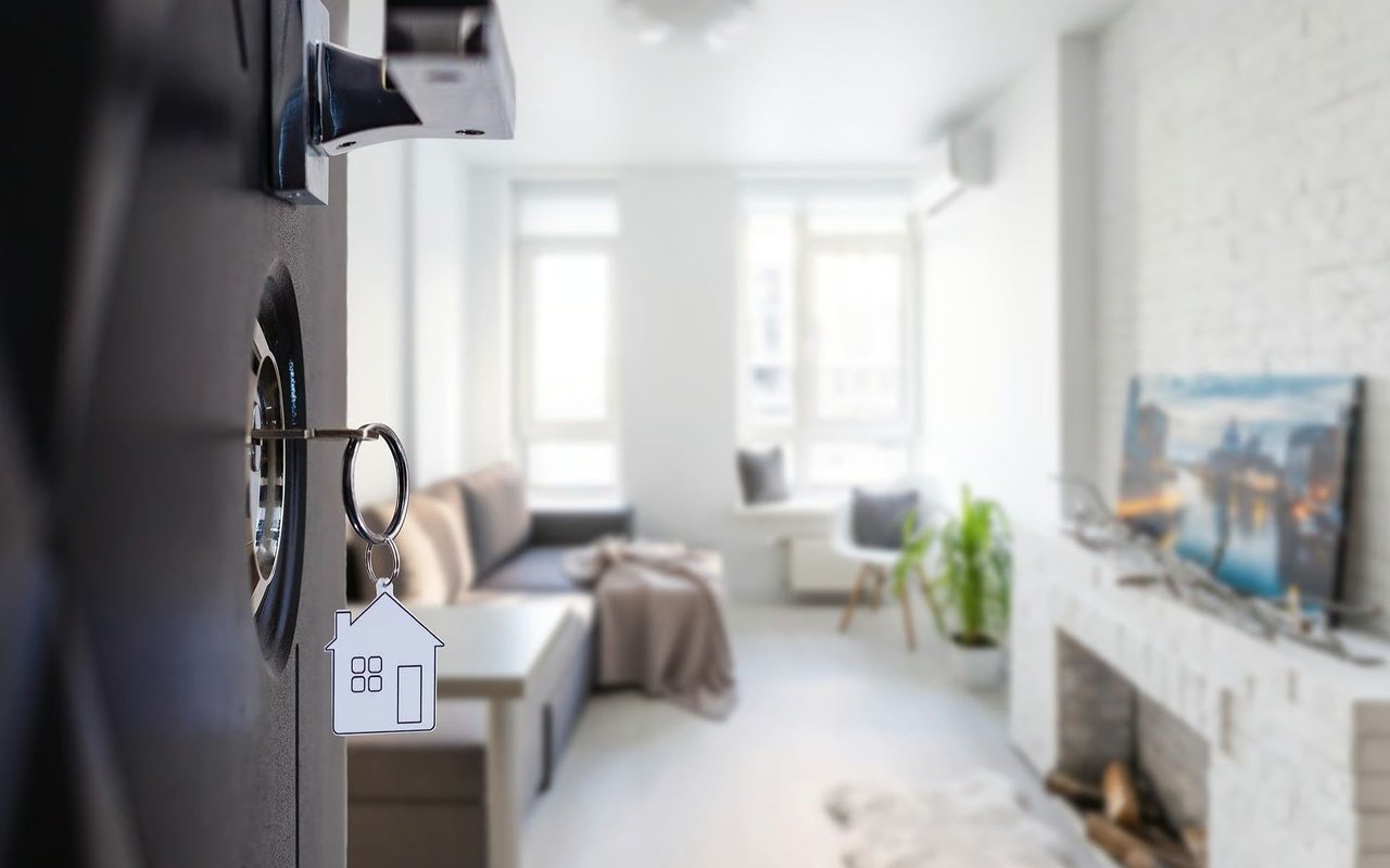 House-shaped keychain hanging from a door lock, with a bright living room in the background.