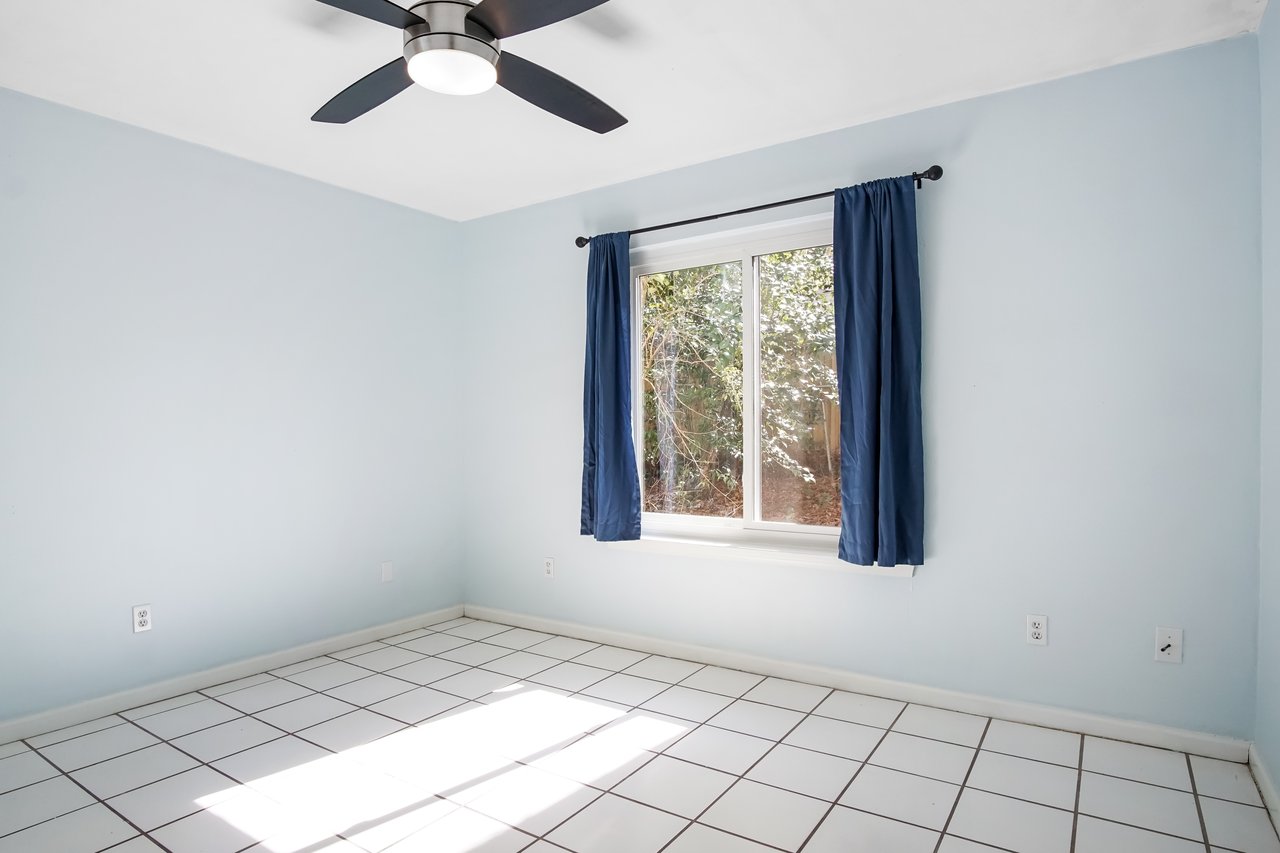 A bright, empty room with light blue walls features a large window with blue curtains, letting in sunlight. The floor is white tile, and a modern ceiling fan hangs above.