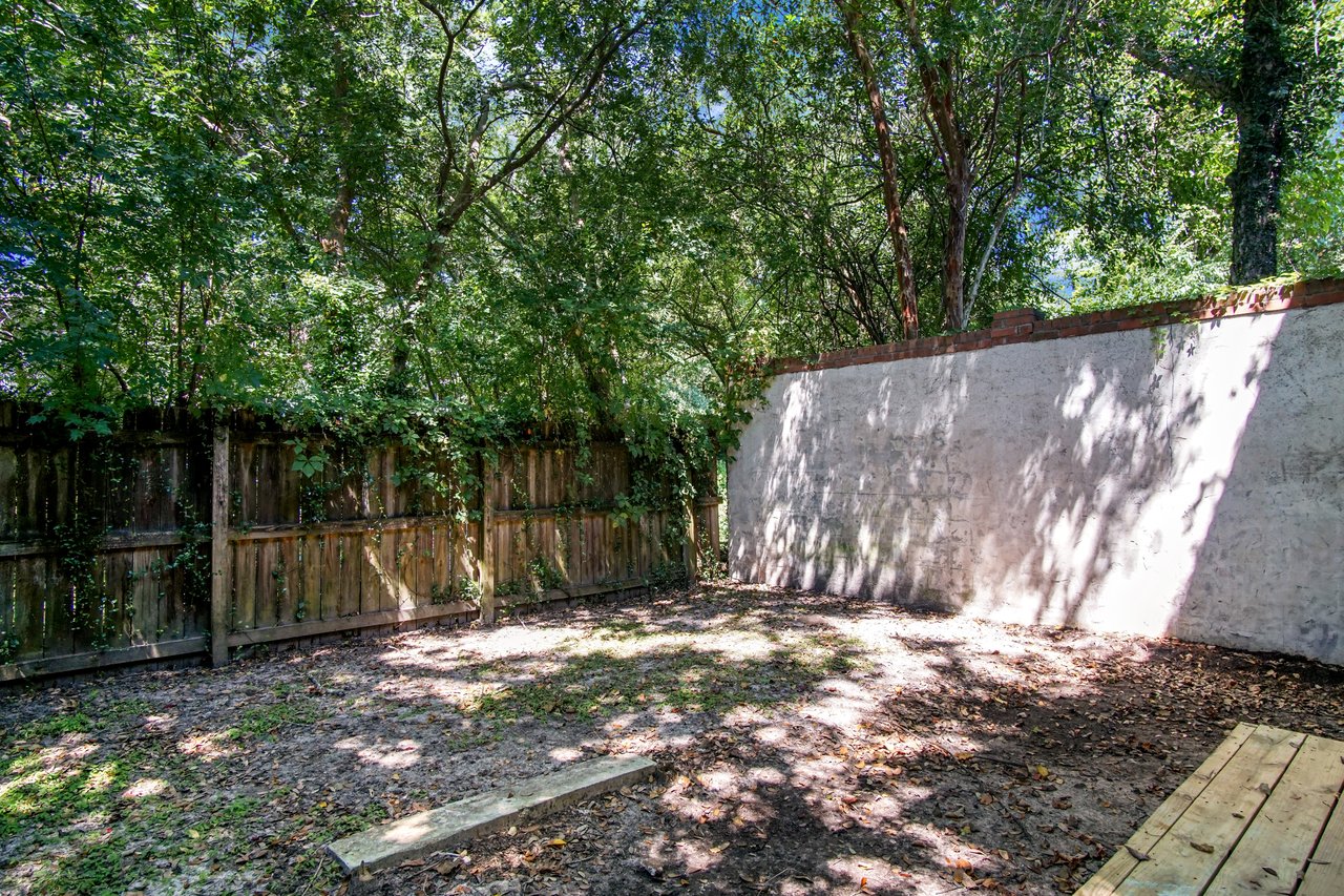 Backyard at 2506 Nugget Lane in Tallahassee, Florida, enclosed with a wood privacy fence and tall stucco wall. The shaded outdoor space features mature trees along the perimeter and a small wooden deck area, offering a private setting.