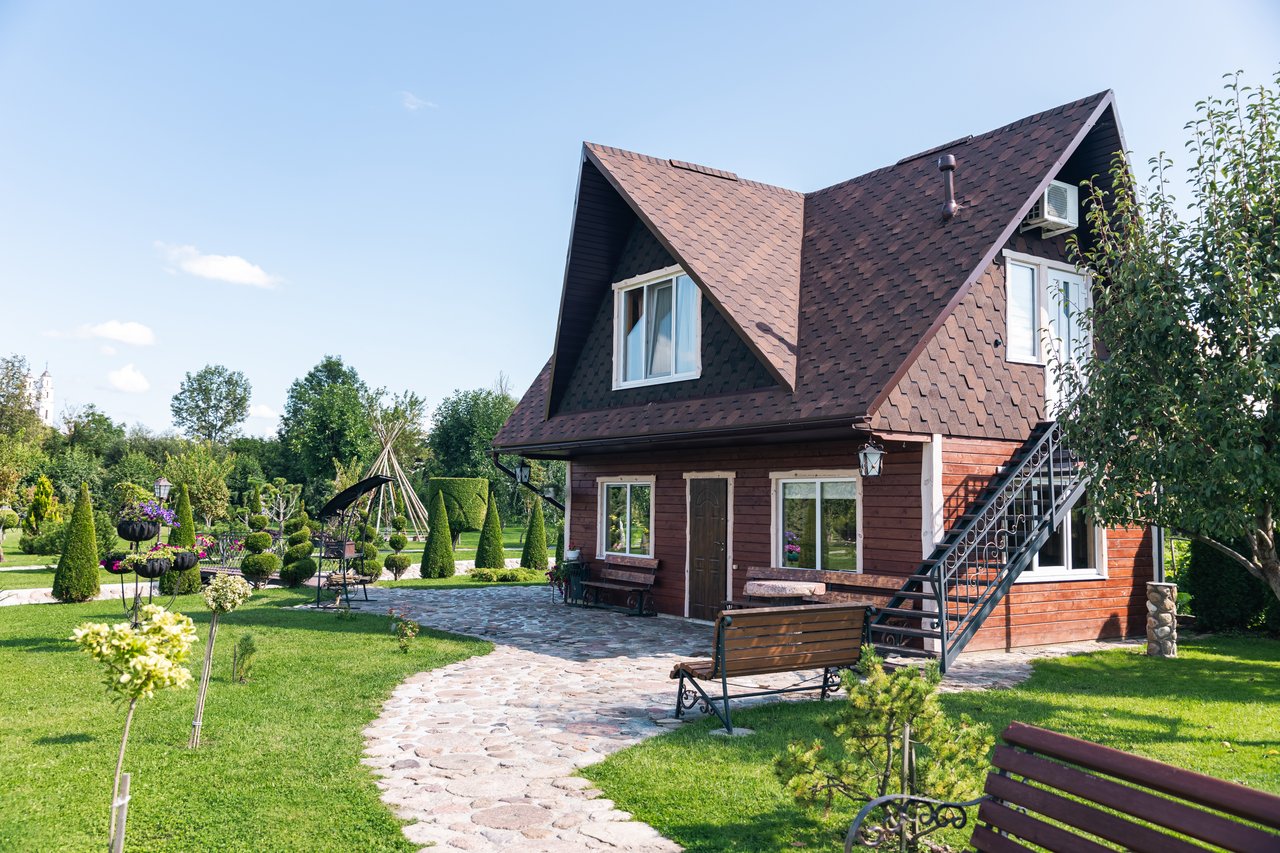 Wooden house with steep shingle roof beside a stone garden path and landscaped lawn.