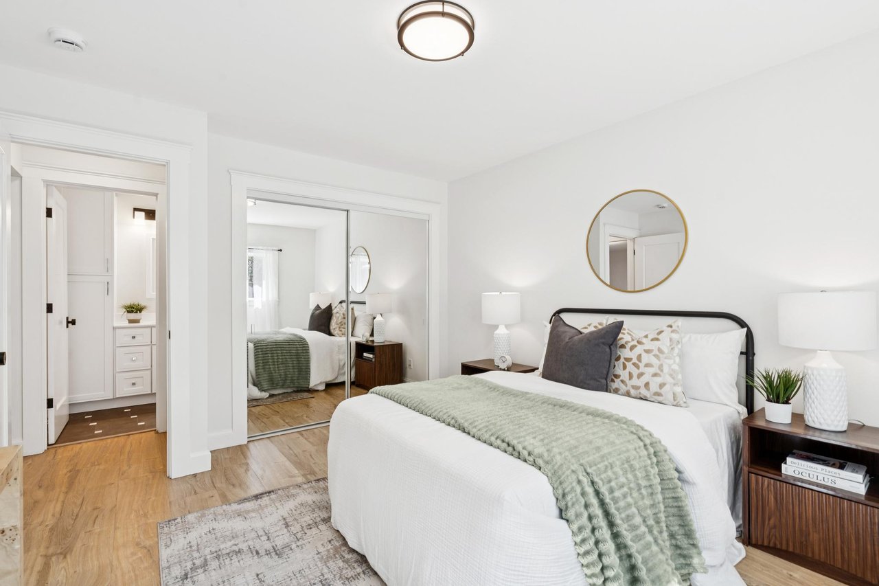 Staged bedroom with a white bed, sage-green throw, round mirror, and mirrored closet doors.