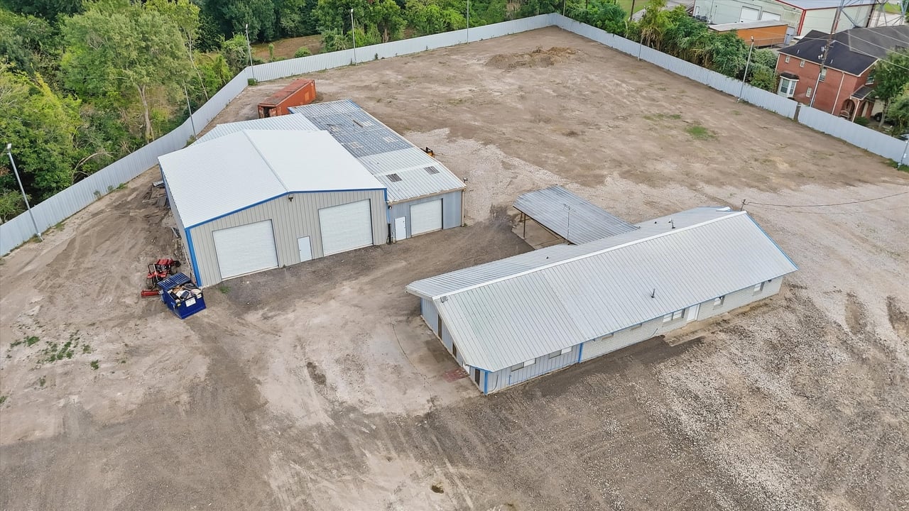 Aerial view of a fenced property with two metal buildings and cleared dirt and gravel.