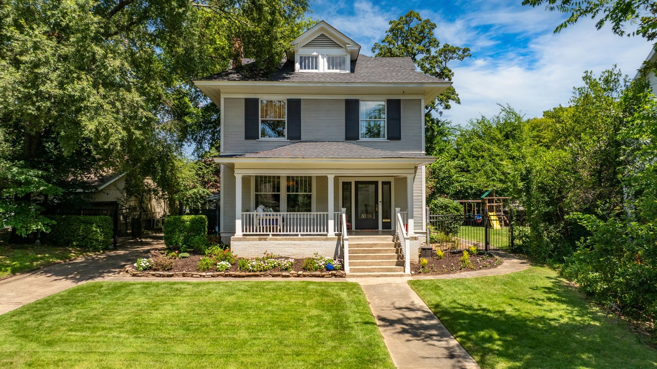 Gray two-story house with a covered porch, landscaped flower bed, and fenced play area.