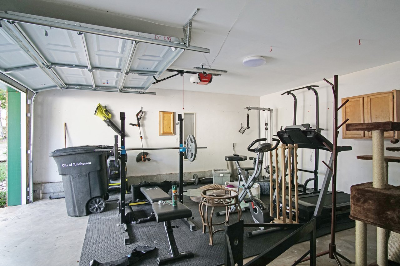 Garage at 529 Collinsford Road, Tallahassee, Florida, currently set up as a home gym. The space includes exercise equipment such as a weight bench, stationary bike, and treadmill. The concrete floor, overhead garage door, mounted storage cabinets, and tools along the wall are also visible.