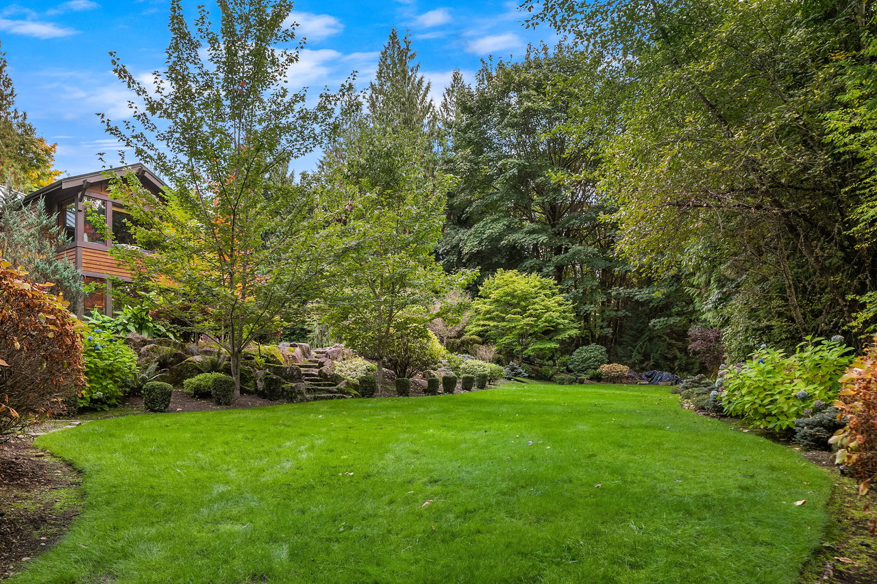 Landscaped home garden with a broad lawn, mature trees, and a partially visible wood-sided house.