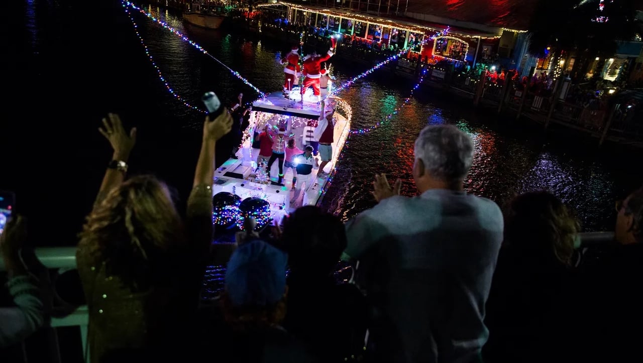 Spectators wave to a Christmas-lit boat during a nighttime waterfront parade.