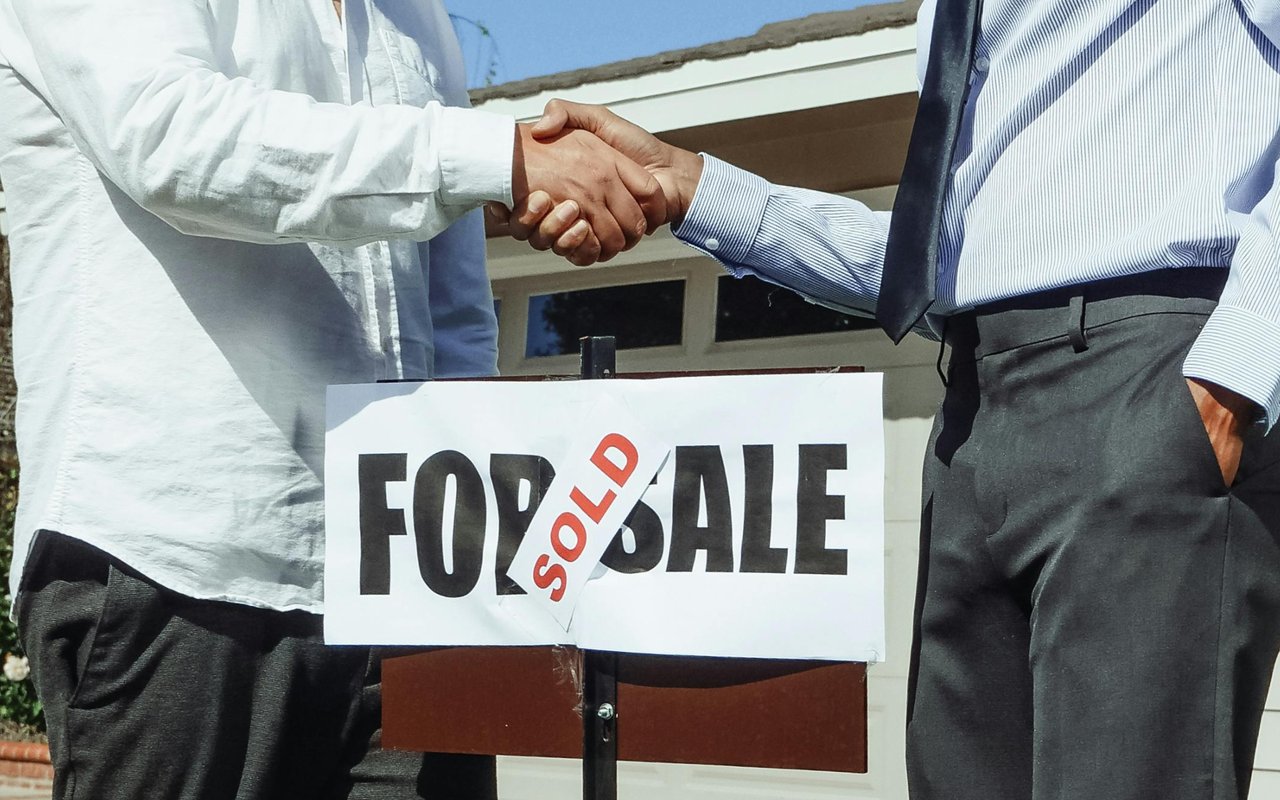 Two people shake hands over a SOLD sign in front of a house.