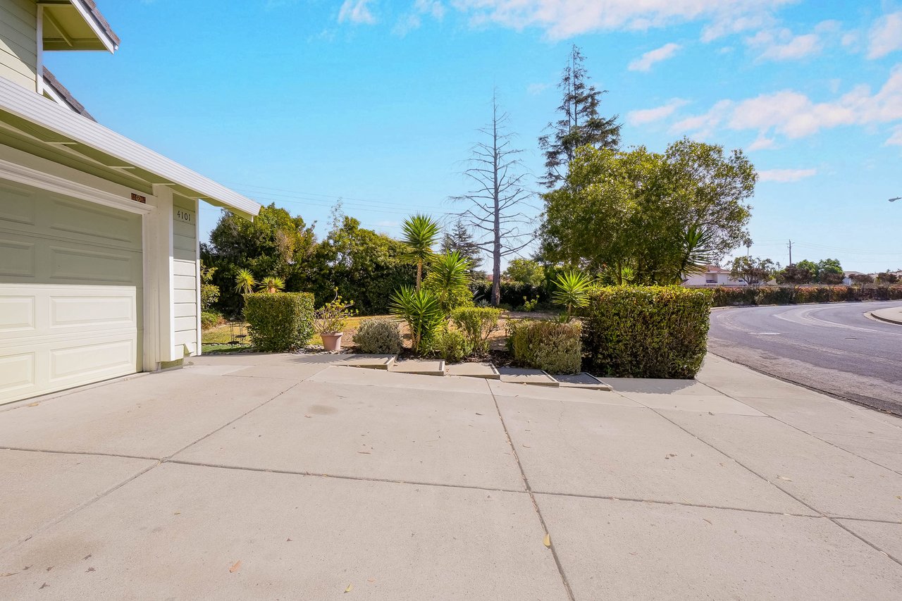 Home exterior with a white garage, wide concrete driveway, and landscaped yard beside a curving street.