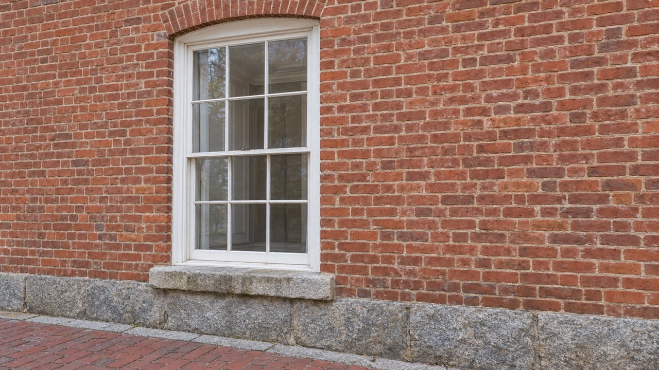 White six-over-six sash window set in weathered red brick above a granite foundation and brick sidewalk.